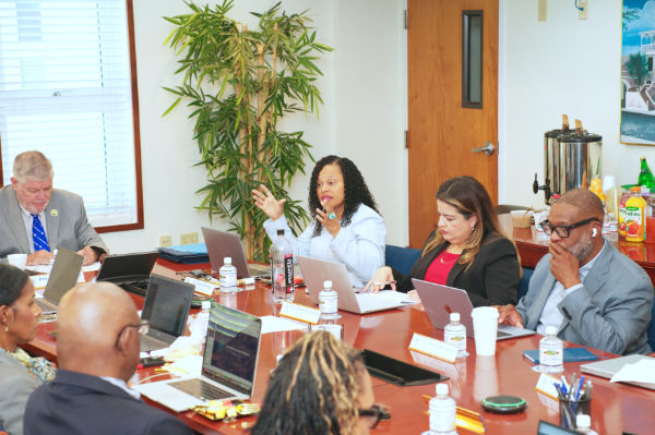 Members of the UVI Board of Trustees seated around a conference table with laptops during a meeting as a speaker gestures while addressing the room.