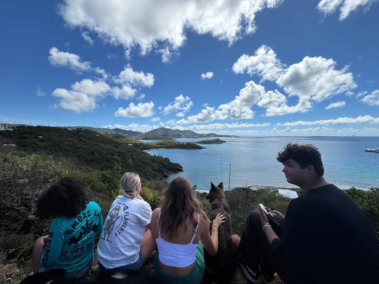 four students and a dog sitting on a hill looking out over a blue ocean.